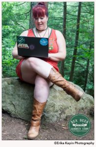 Heather Sitting on a rock in the forrest working on a laptop. They are wearing an orange dress, and light brown cowboy boots 