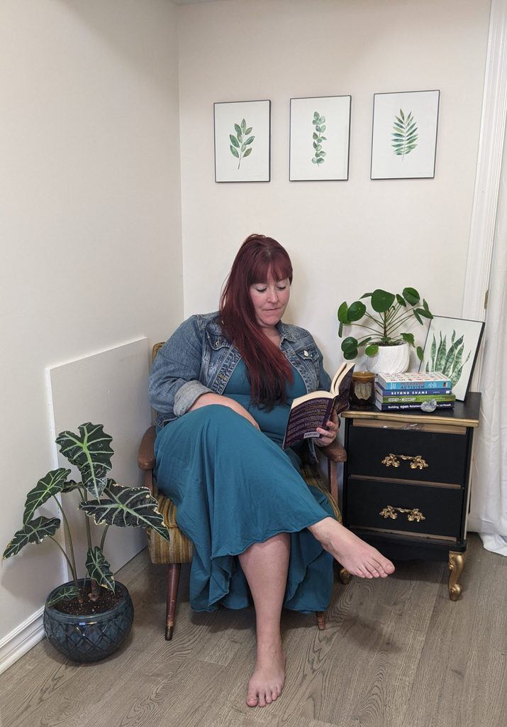 a white femme with red hair in a long blue dress and seat jackets sits reading a book among a number of plants.