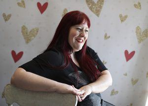 Heather sits sideways behind a while chair, laughing. The background is pink with red and gold hearts.